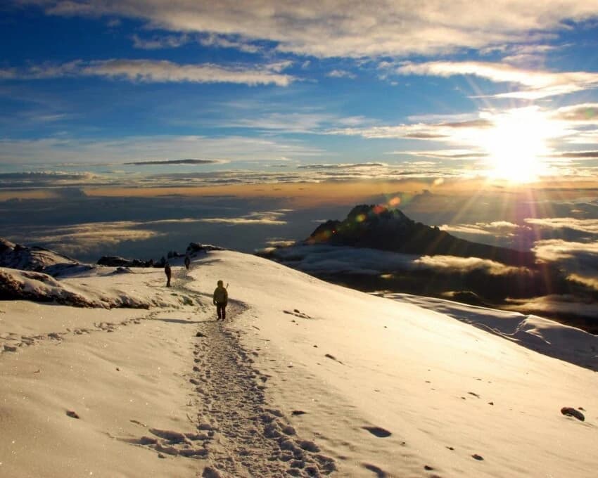 Tiny hiker on a snowy Kilimanjaro summit trail at sunrise above clouds