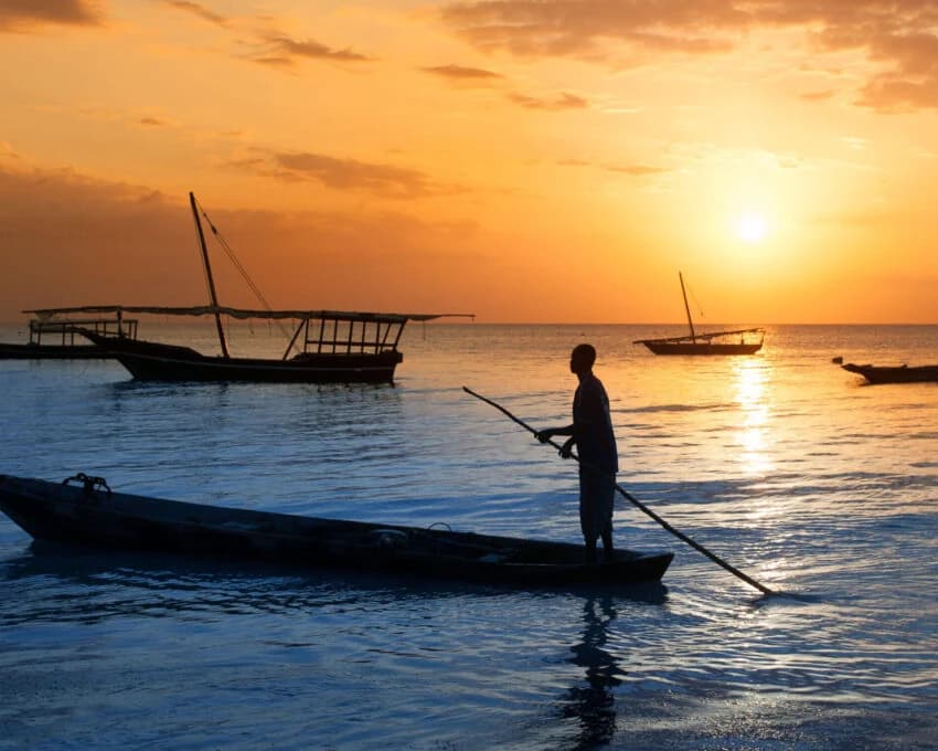 Fisherman poling a canoe past dhows at sunset