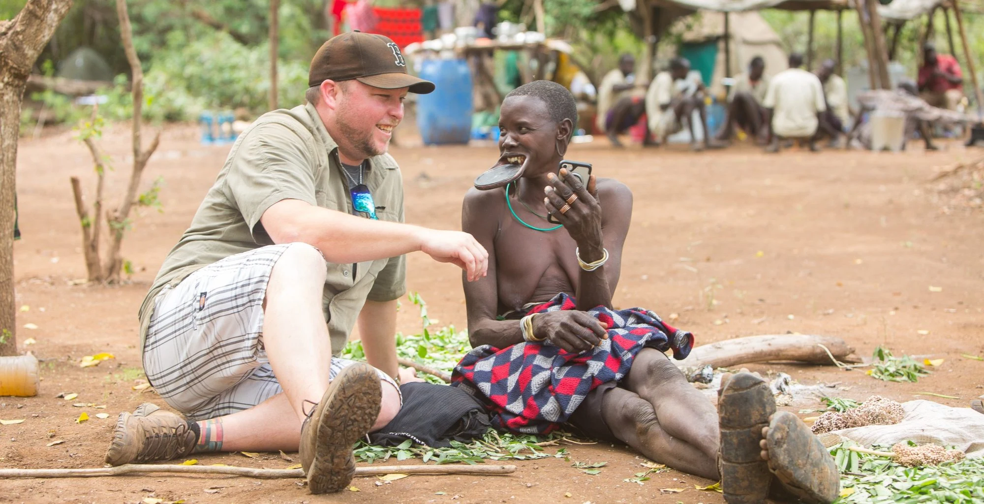 A visitor sits and talks with a Mursi man in the shade of trees.