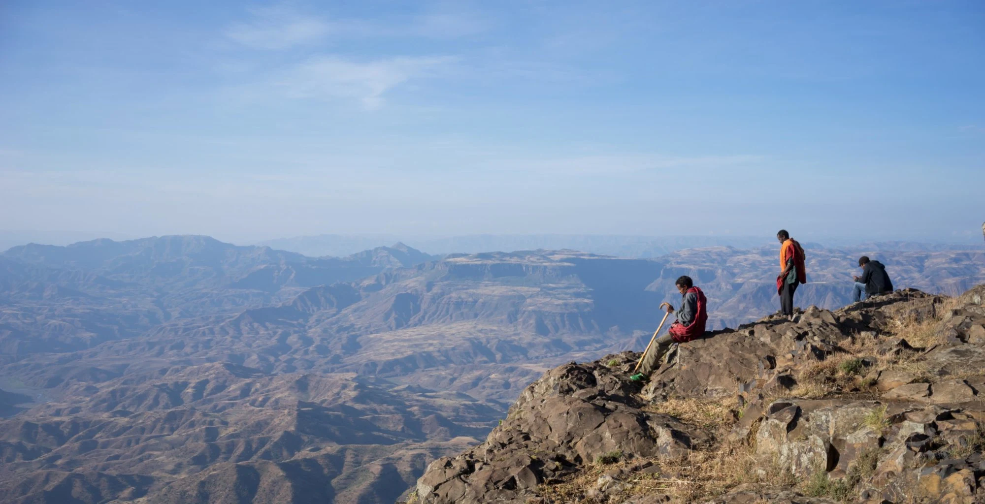 Three walkers rest on a rocky outcrop above wide Tigray valleys.