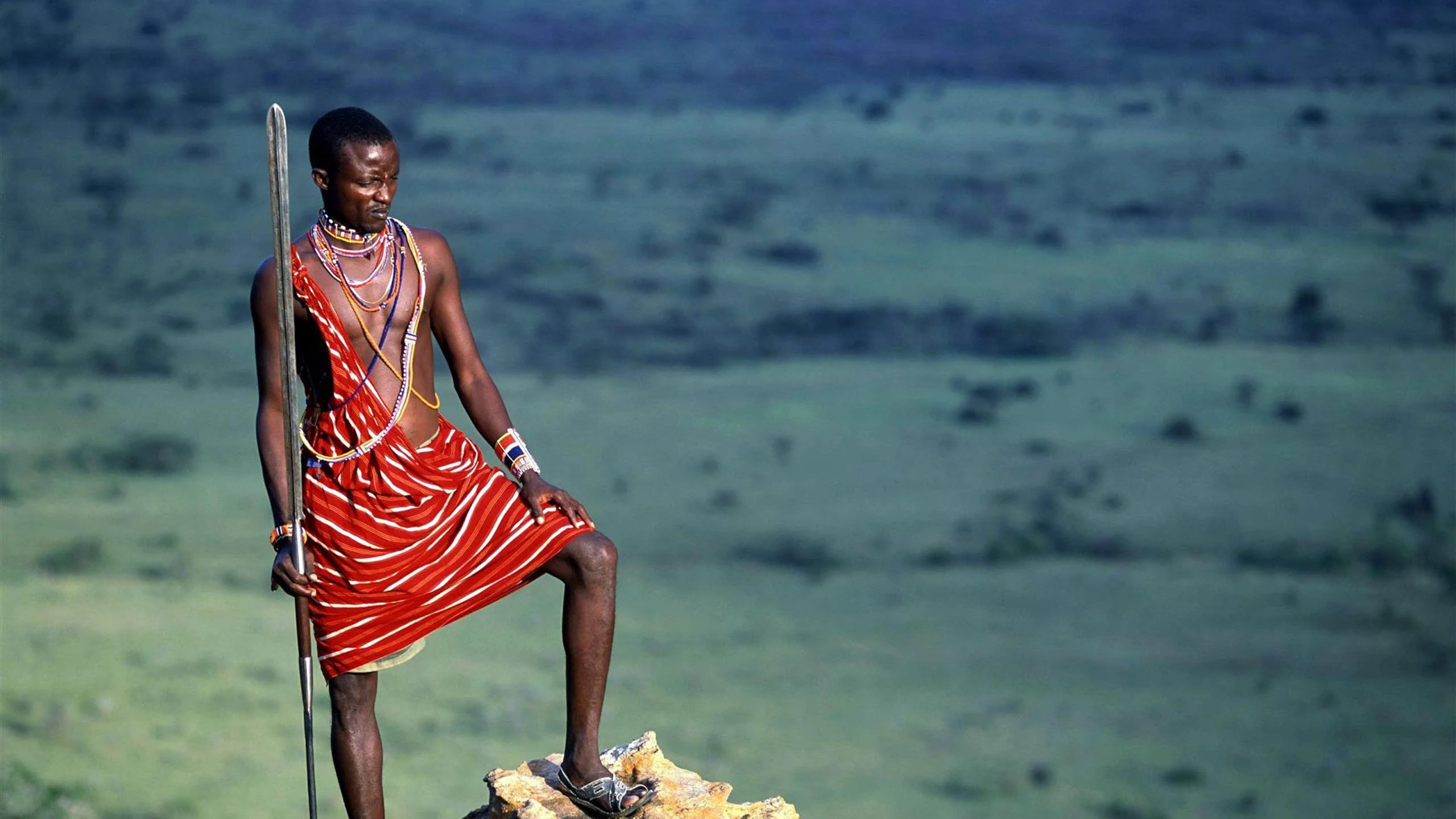 A Maasai man in a red shuka standing on a rock above the plains in the Chyulu Hills
