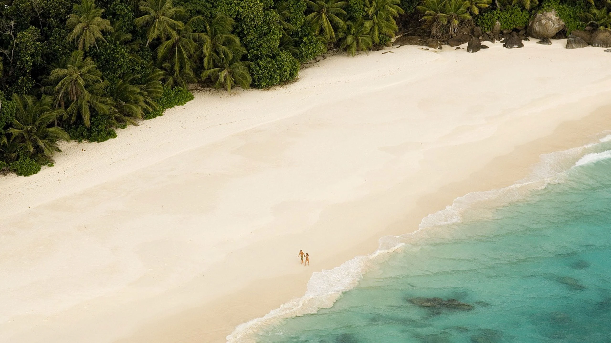 Aerial of an empty white beach with two tiny walkers and forest edge