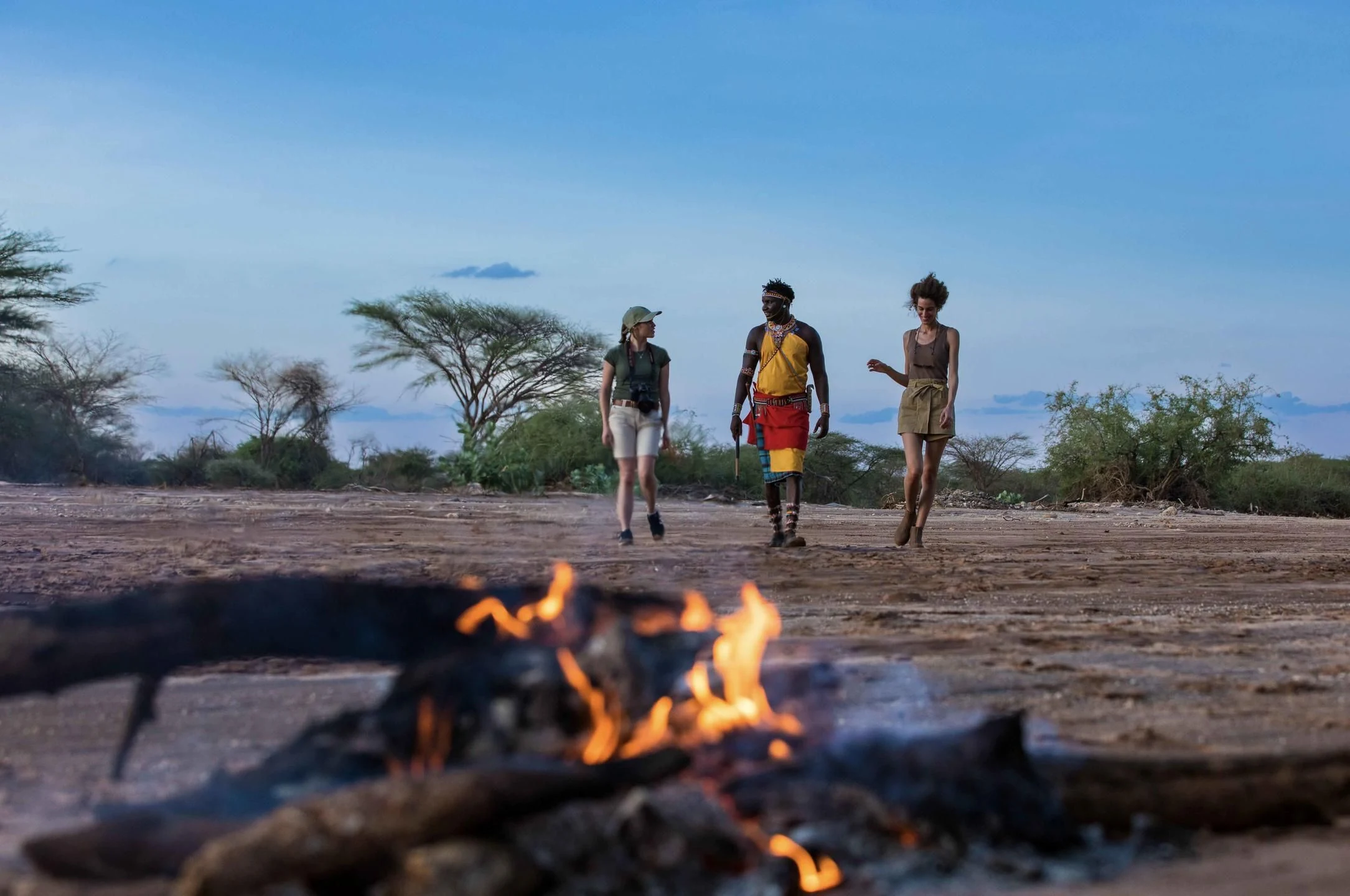 Three walkers crossing dry riverbed at dusk past a campfire