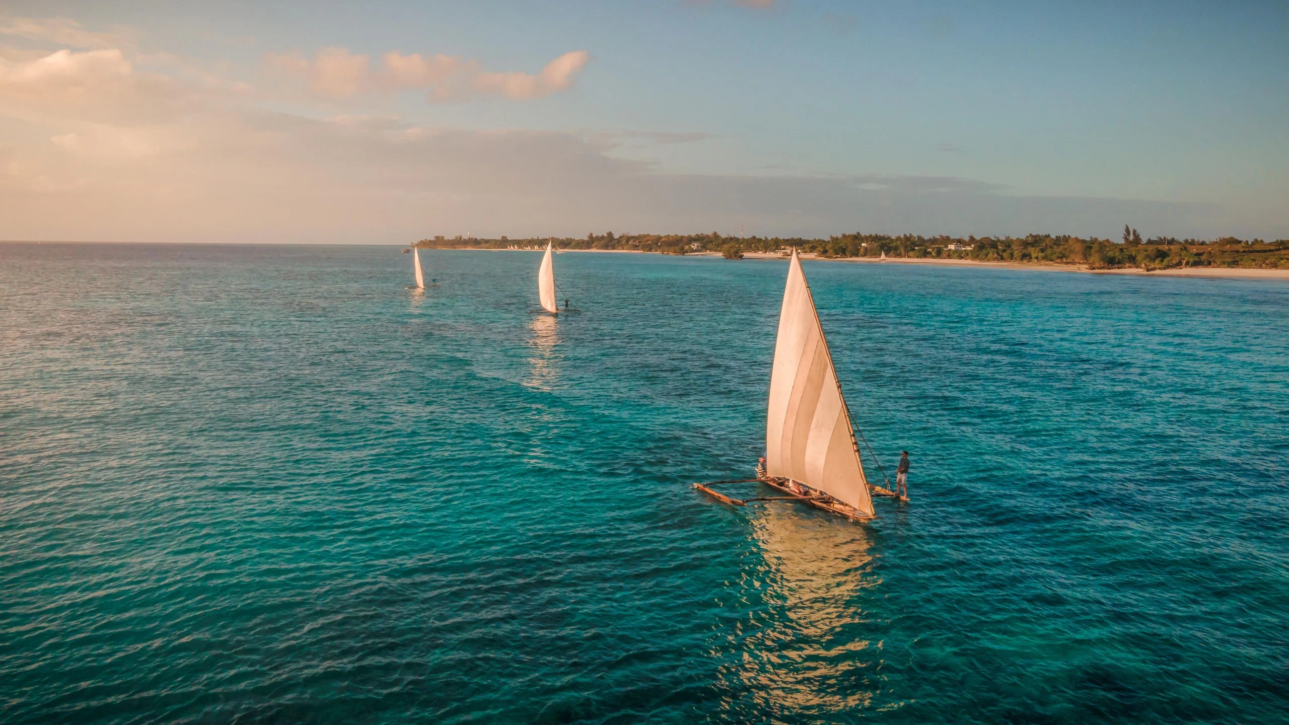 Three traditional sailing boats heading out across shallow turquoise water