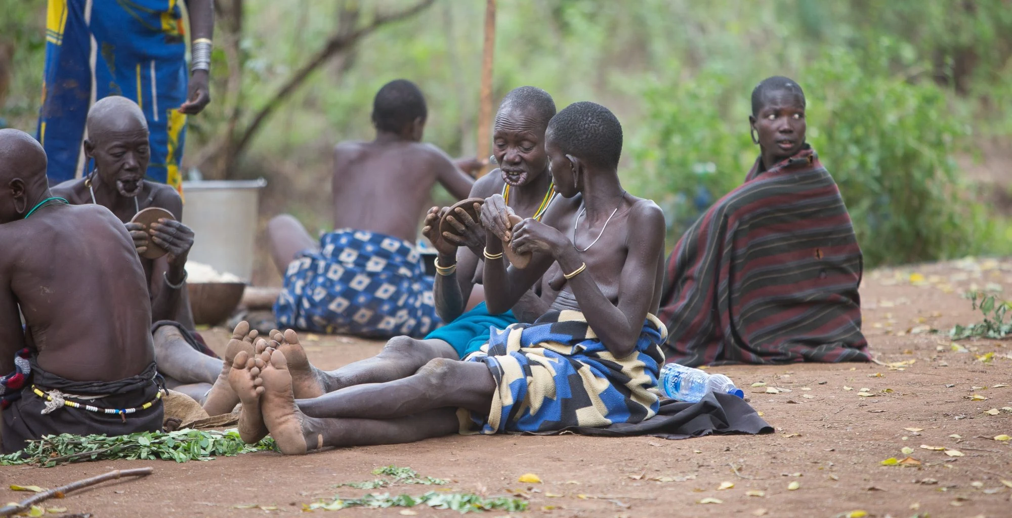 Mursi men and women sit together on the ground beneath trees.