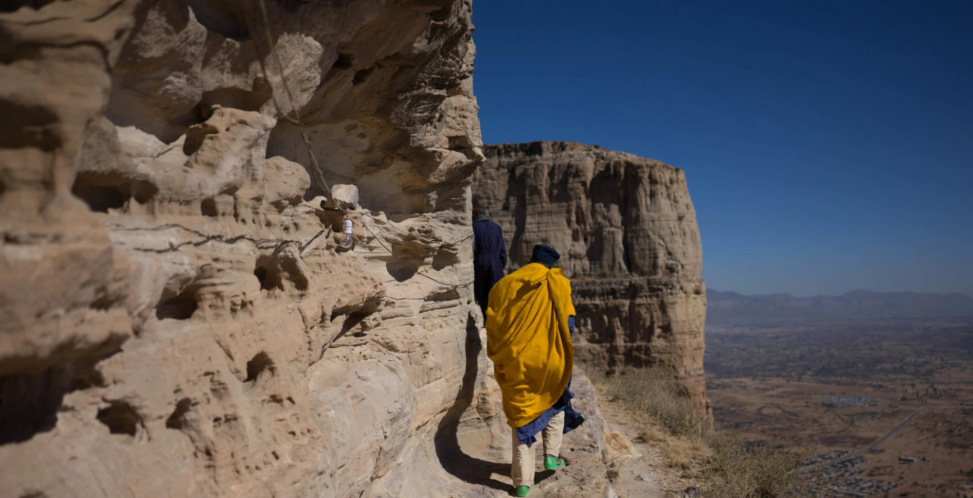 A person in a yellow robe walks a rock ledge high above a valley near Lalibela.