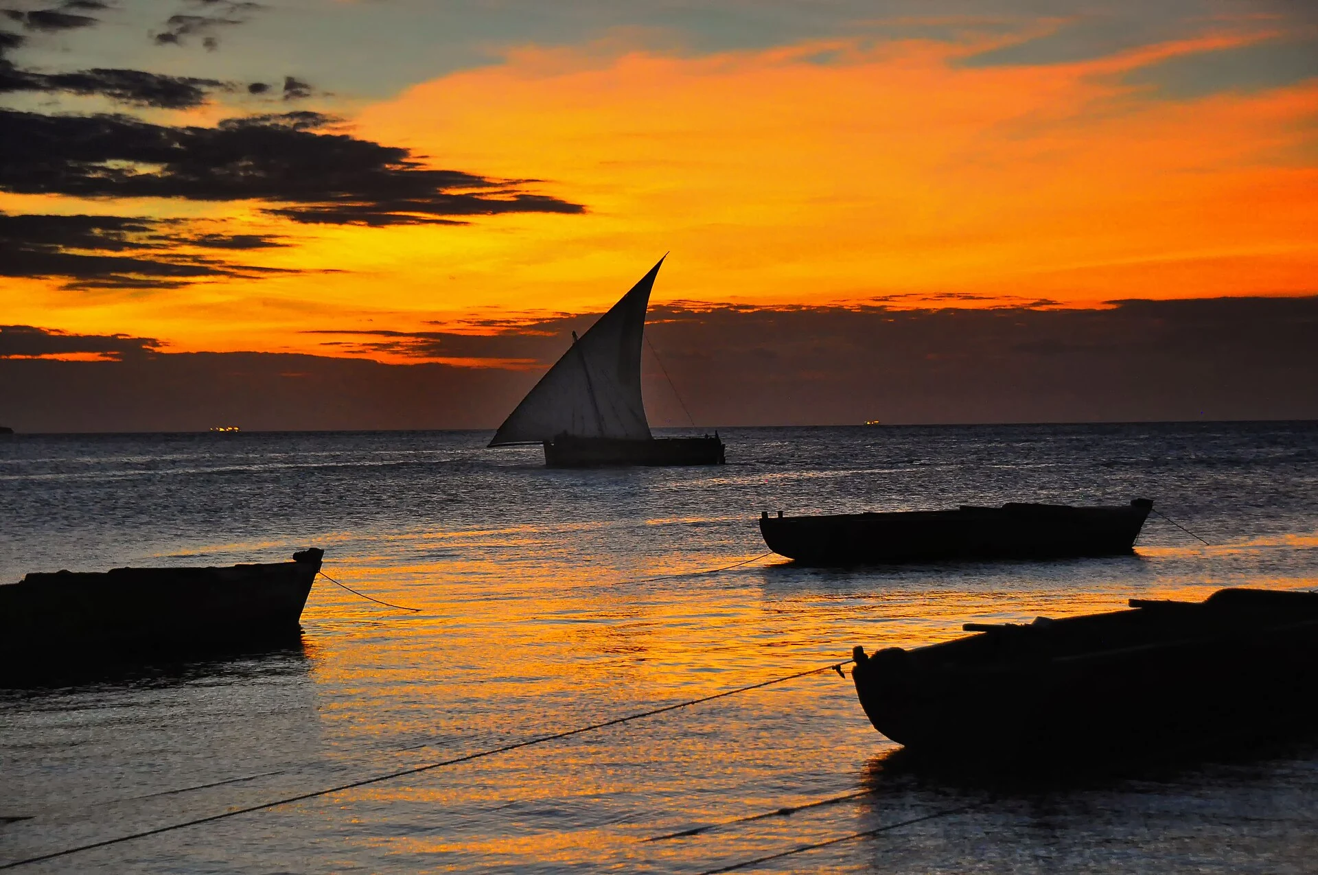 A dhow sailing past moored boats under an orange Zanzibar sunset