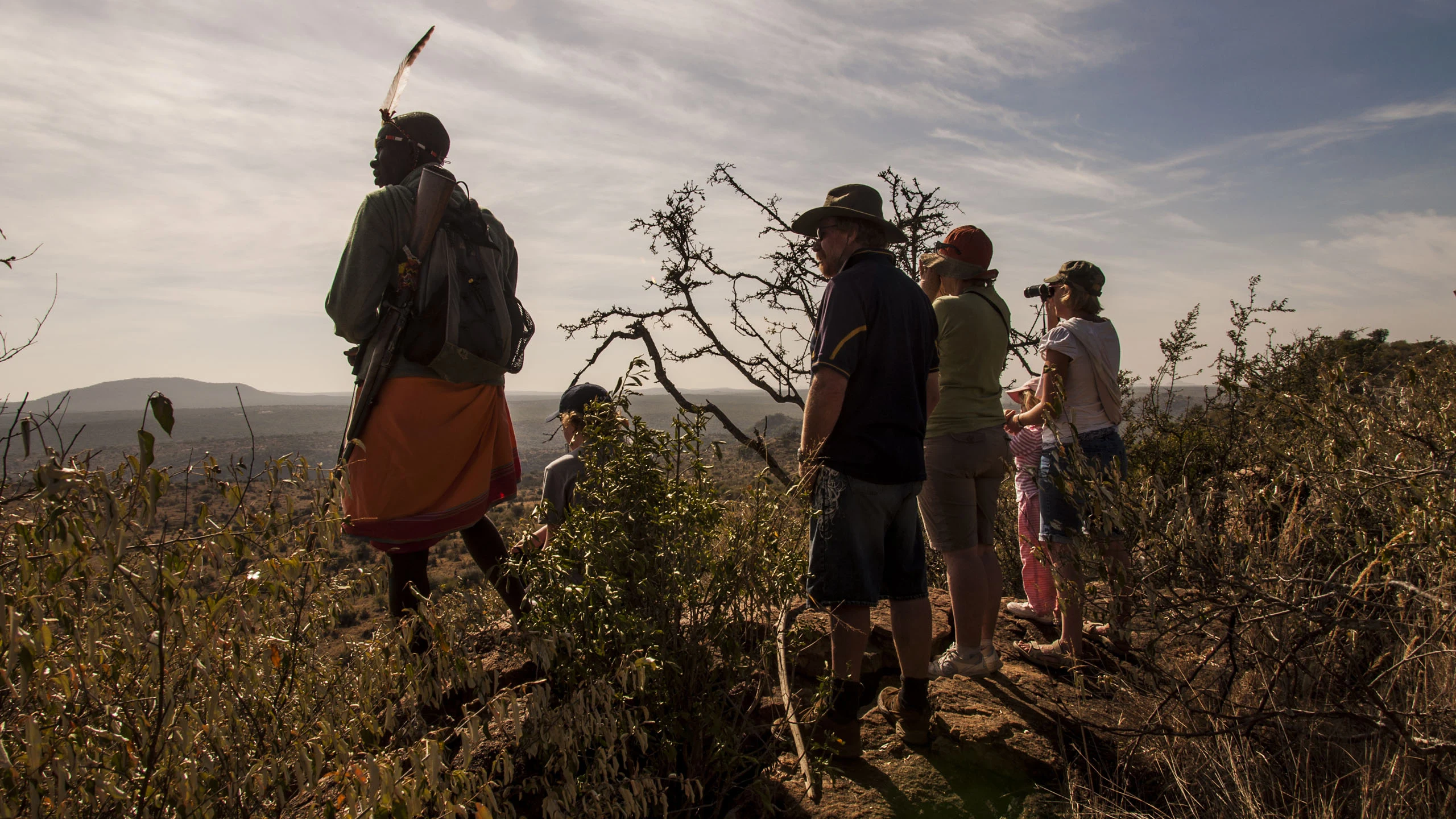 A scout with a rifle on his back stands on a ridge with walking guests, who scan the Laikipia bush through binoculars