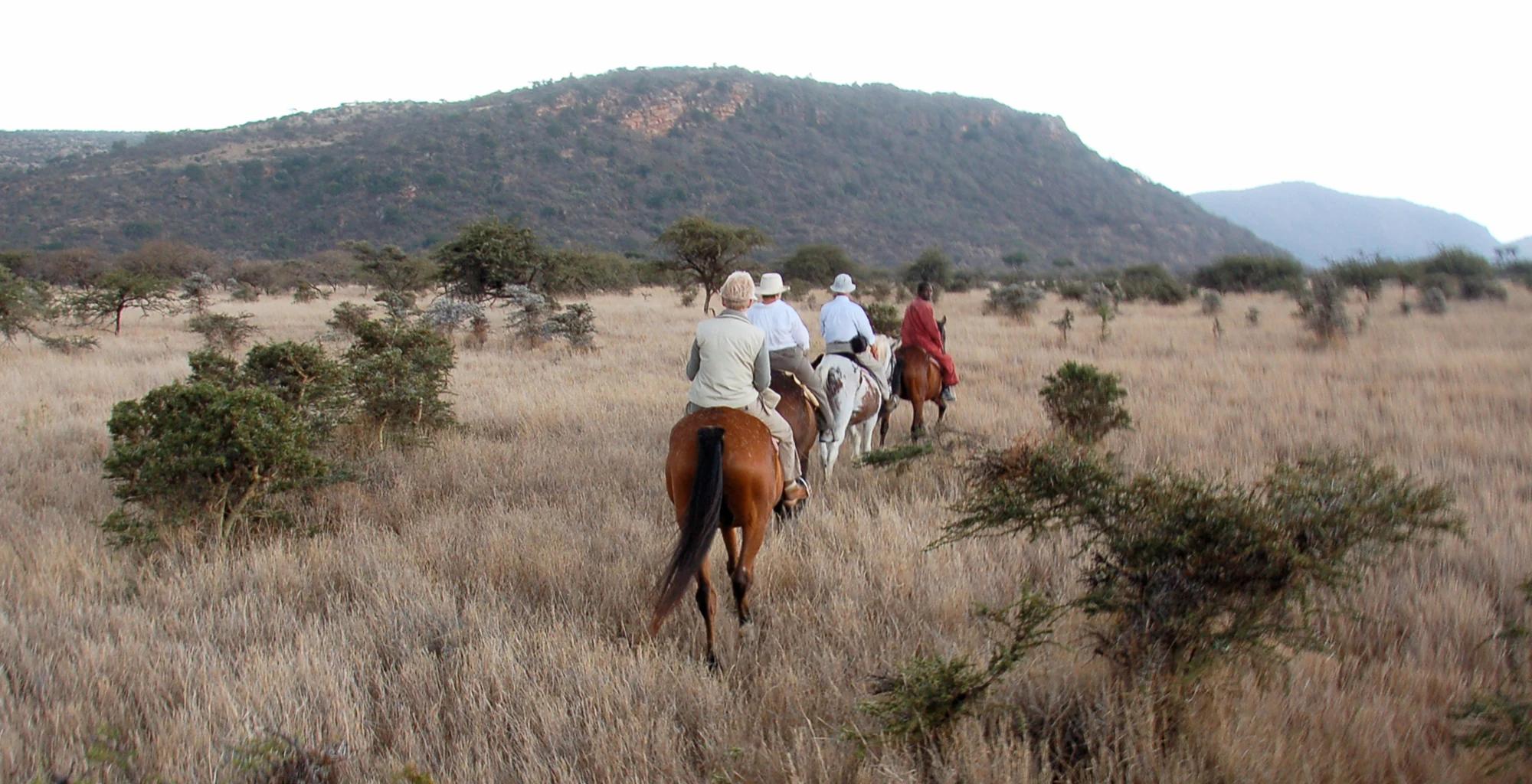 Riders cross open scrubland on horseback beneath a rocky hill