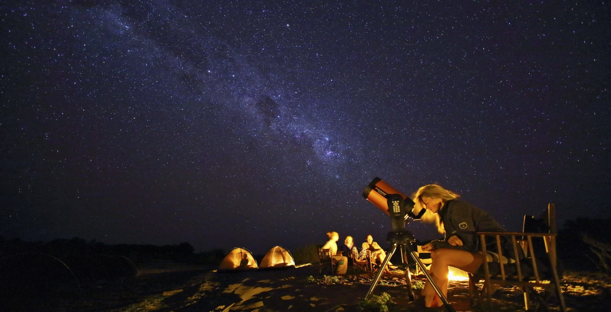 People with a telescope under the Milky Way beside a small camp