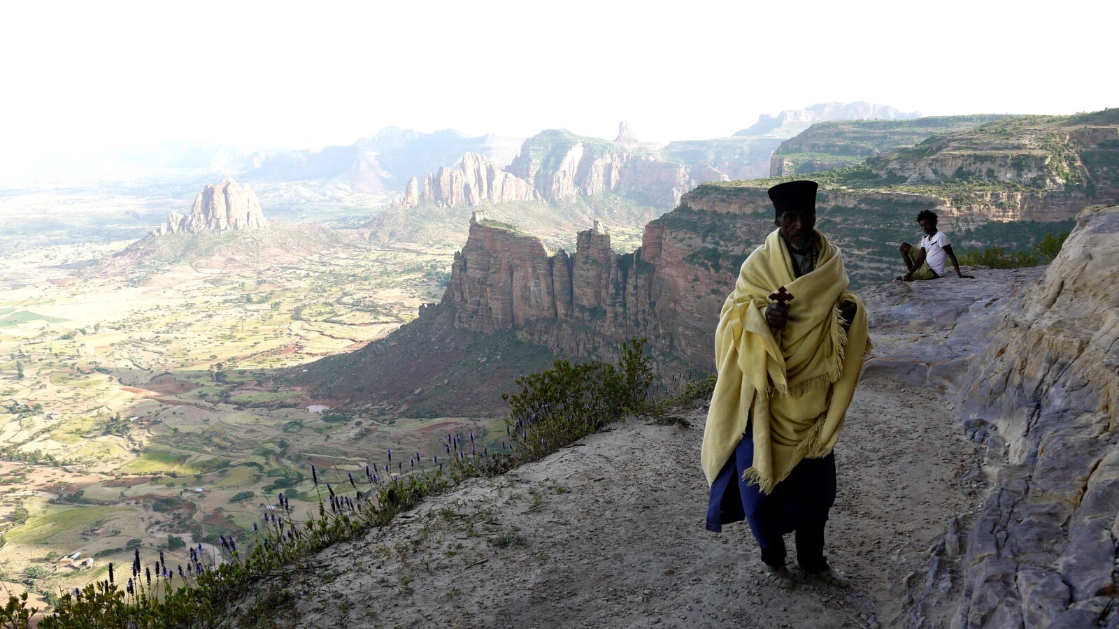 A man in a shawl stands on a cliff edge above a deep Simien valley.