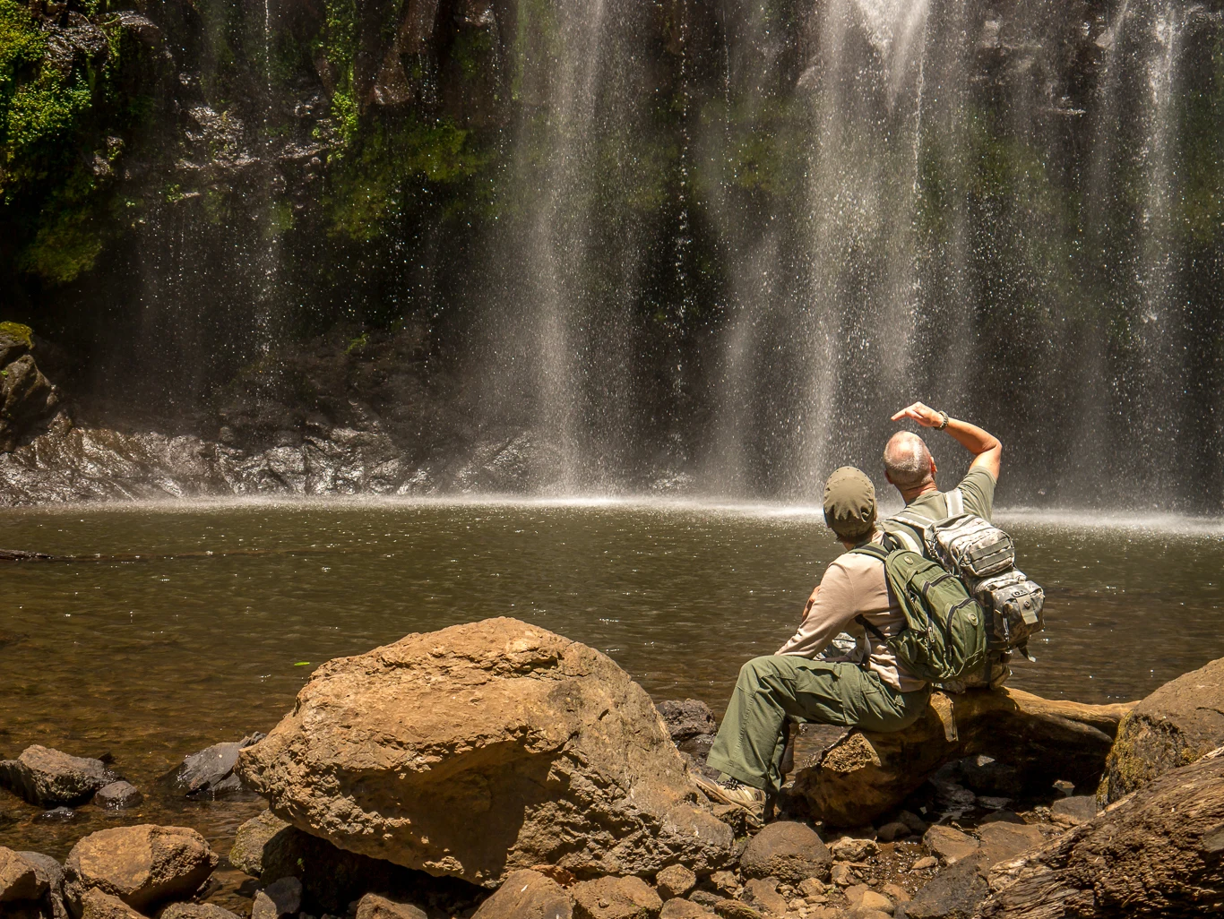 Two hikers resting on rocks at the foot of Materuni waterfall