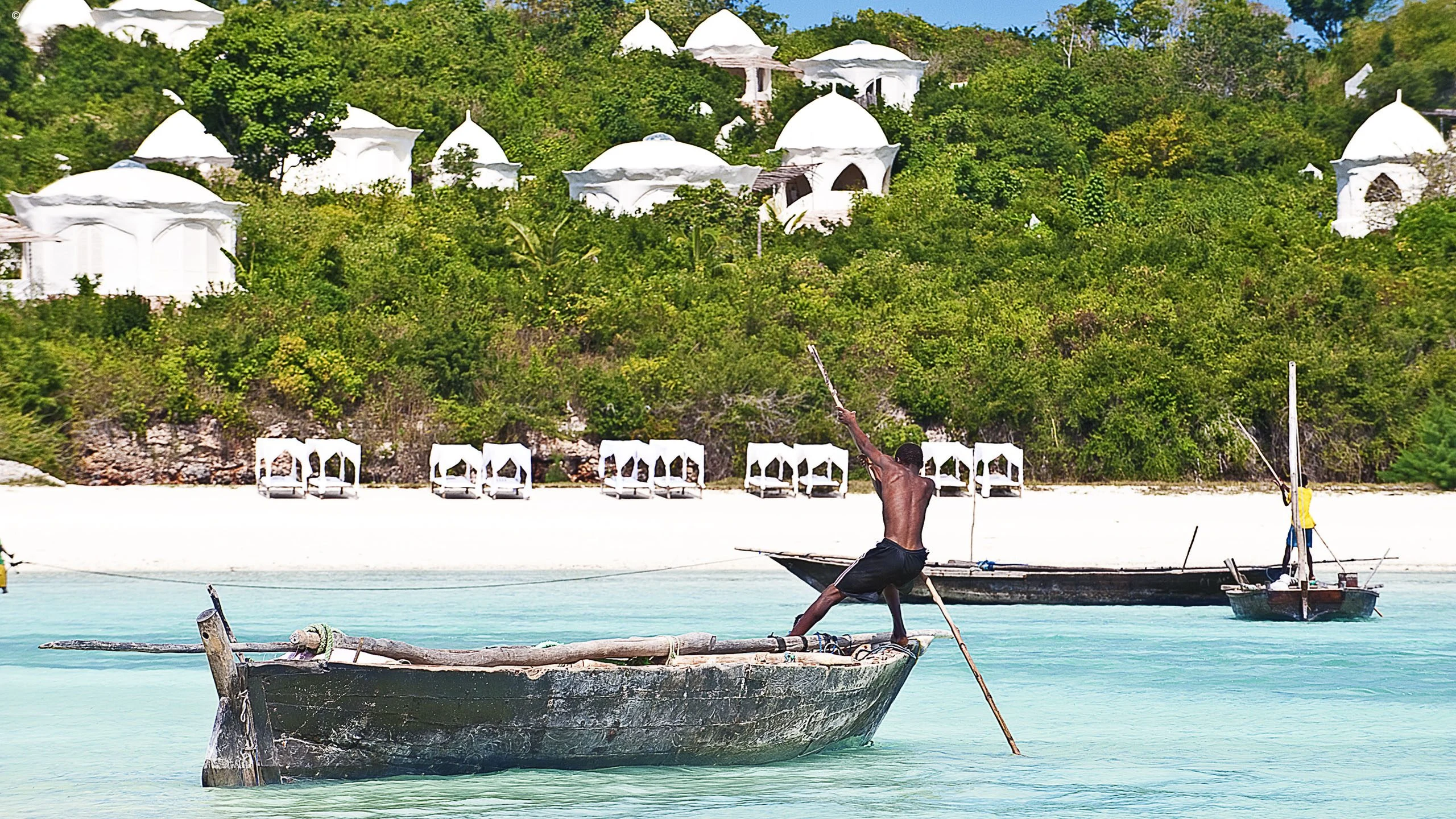 Boatman poling a wooden boat in turquoise water, white villas on the hill