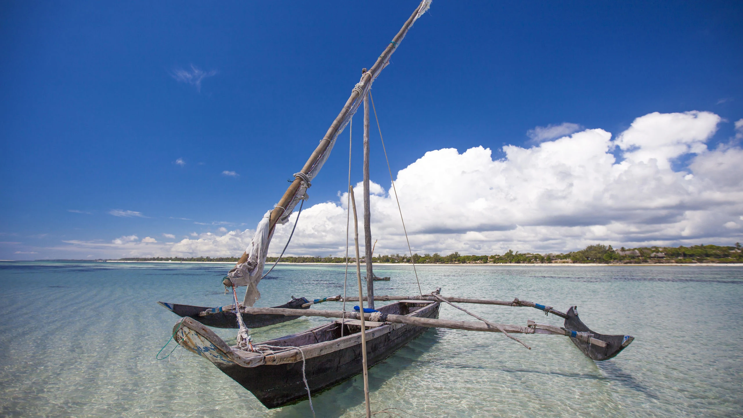 A wooden outrigger sailing boat, its sail furled, lies at anchor in clear turquoise shallows off a low wooded shore on the Kenyan coast