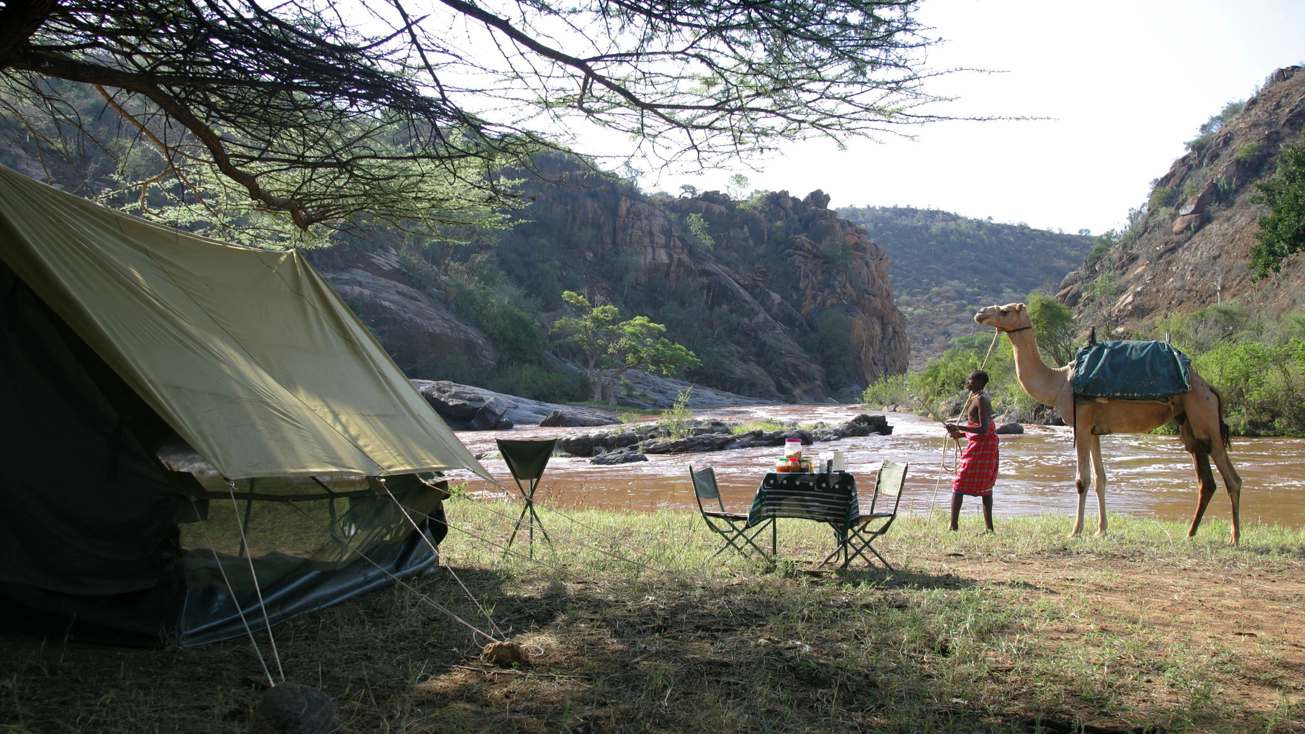 A man in red cloth holds a saddled camel beside a riverside camp with a canvas tent, a small table and folding chairs, rocky hills behind