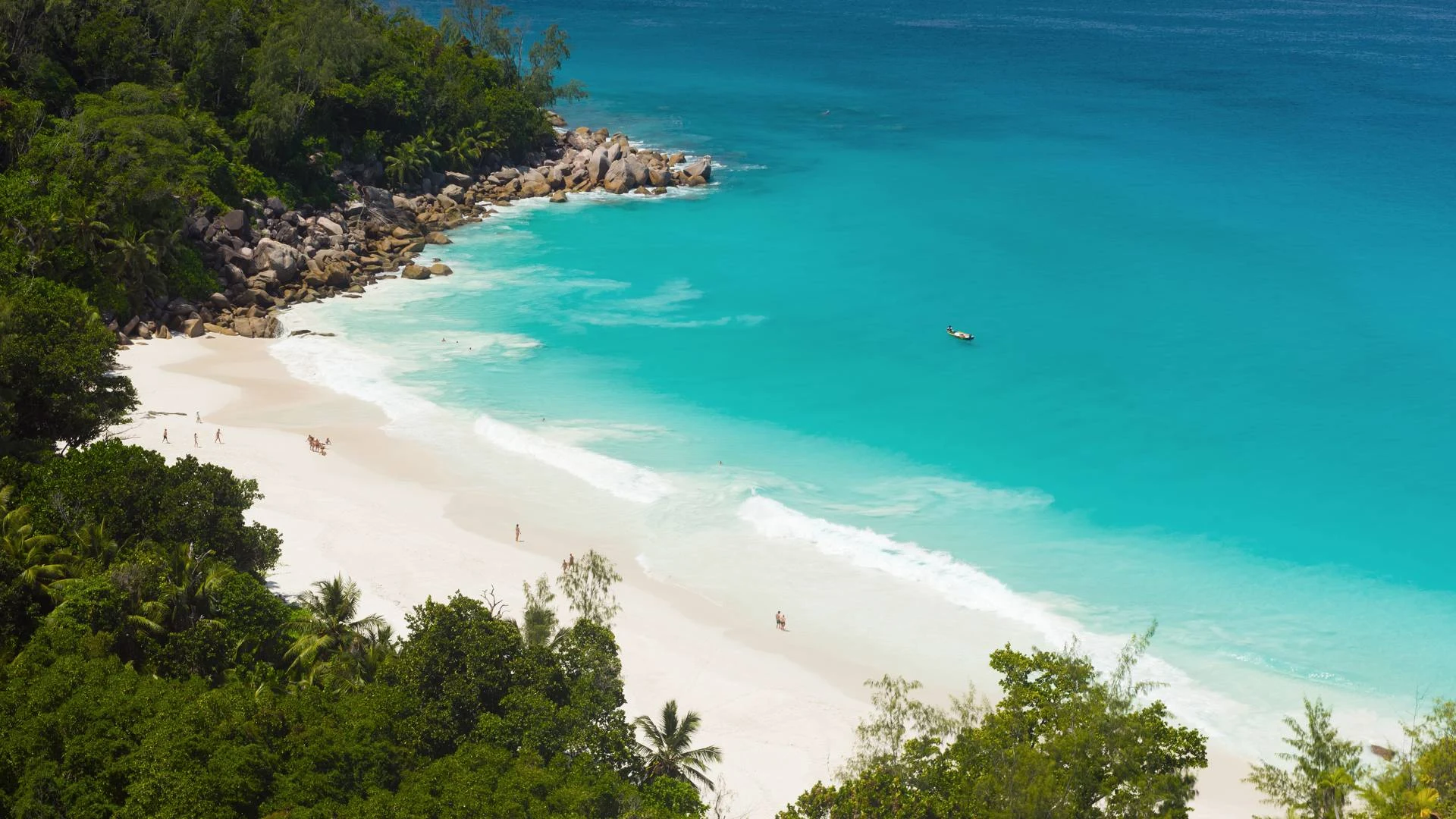 A white beach curving around a turquoise bay below forested hills, seen from the air