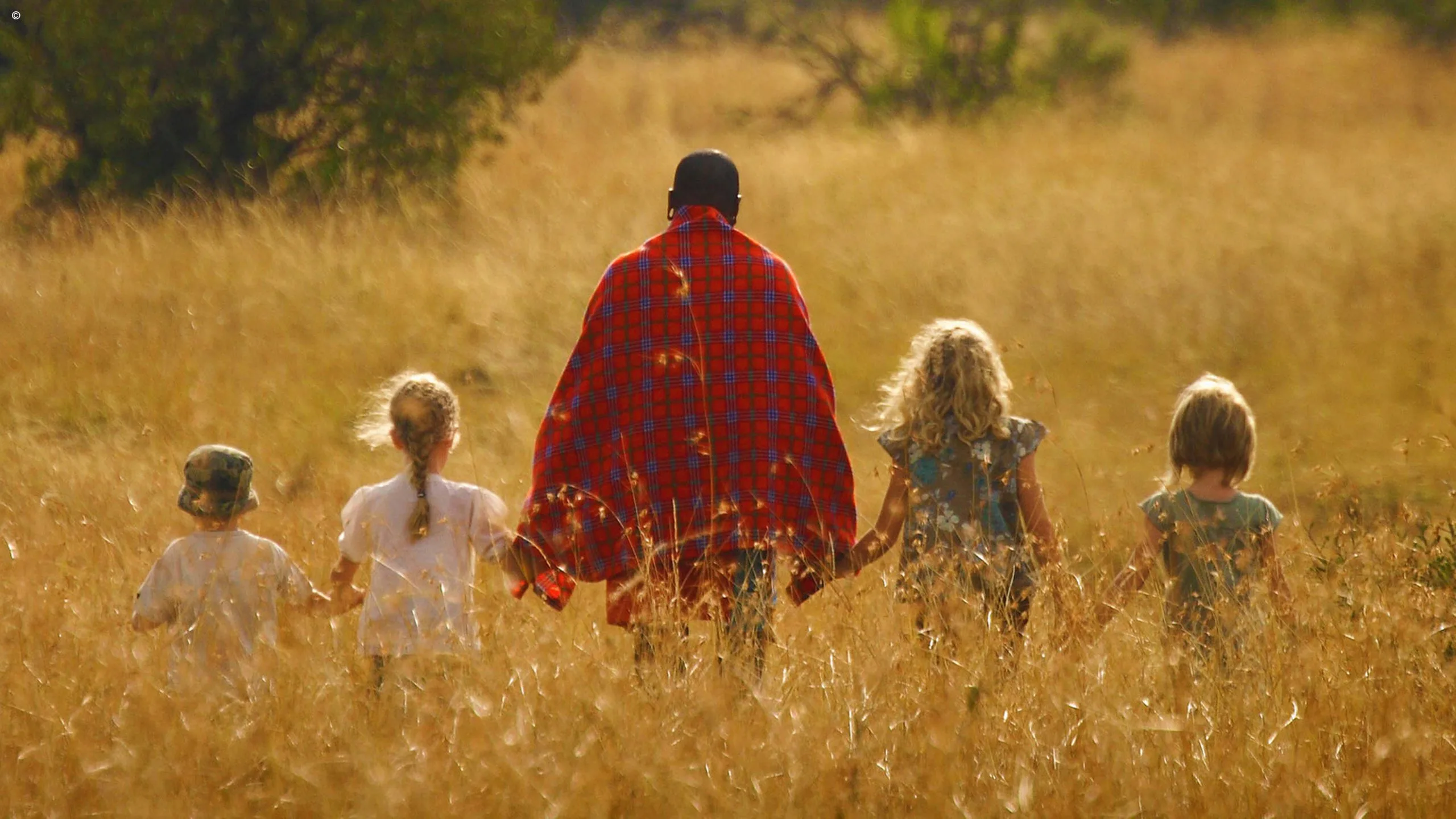 A guide in a red shuka walks hand in hand with four children through golden grass
