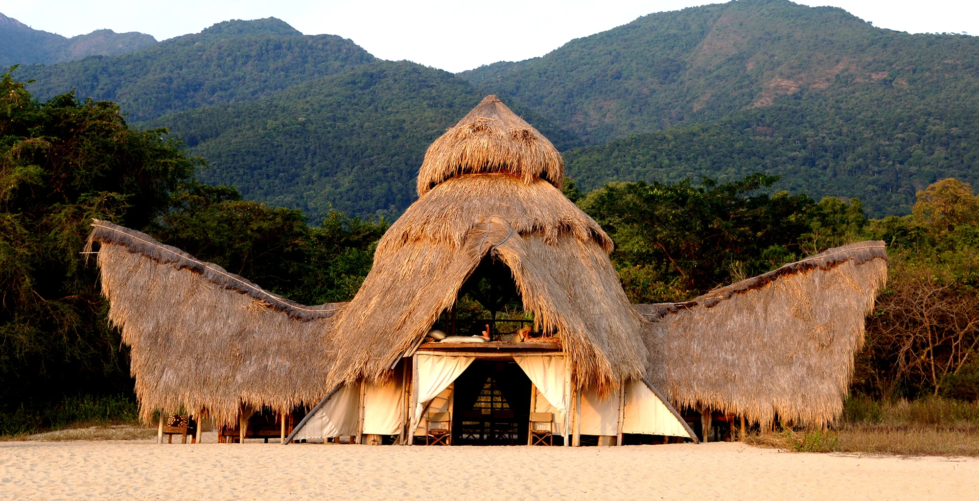 Winged thatched lodge on the beach below the forested Mahale Mountains, Greystoke Mahale