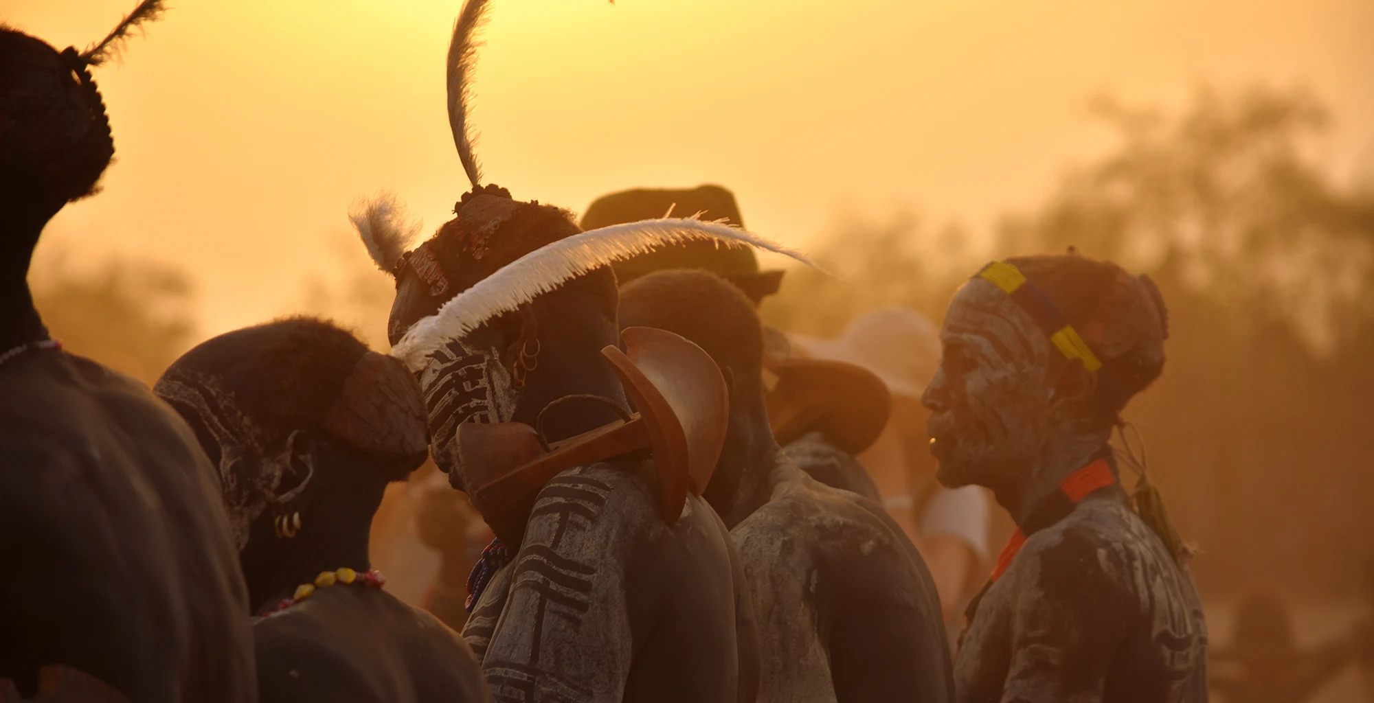 People in feathered headdresses silhouetted against a golden Omo Valley sunset.