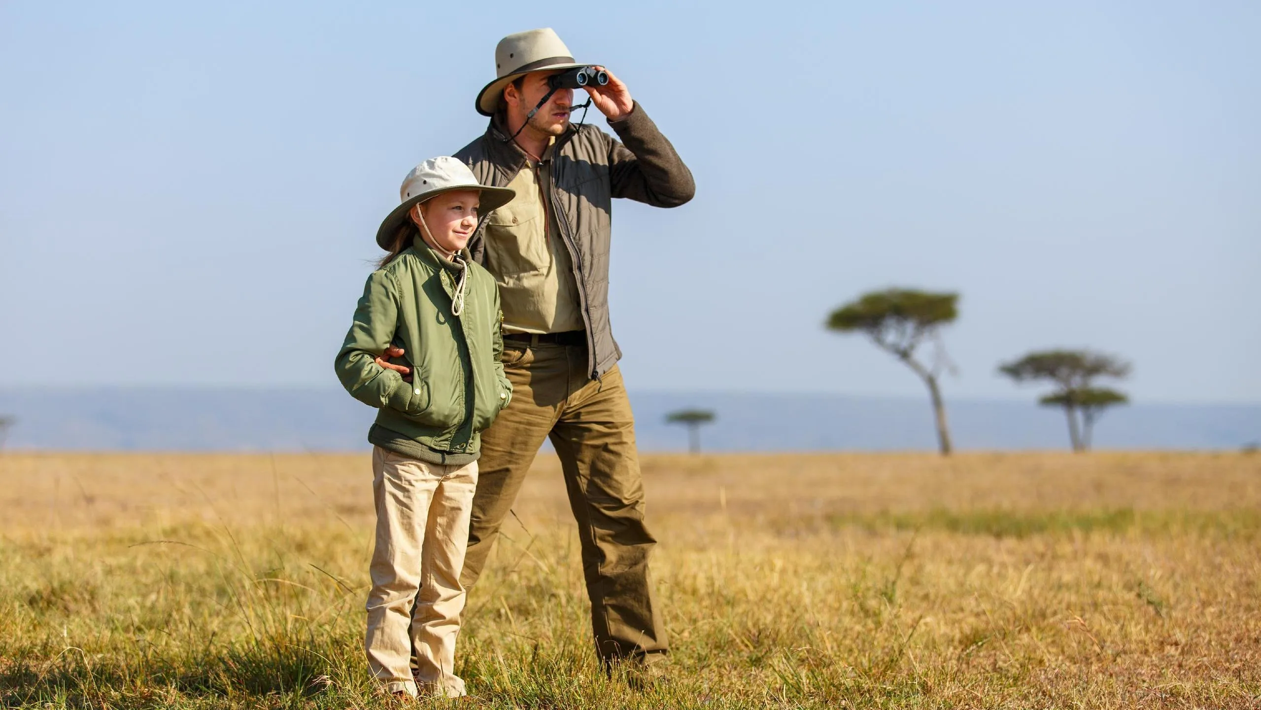 Parent and child in safari hats watching wildlife across open Maasai Mara grassland