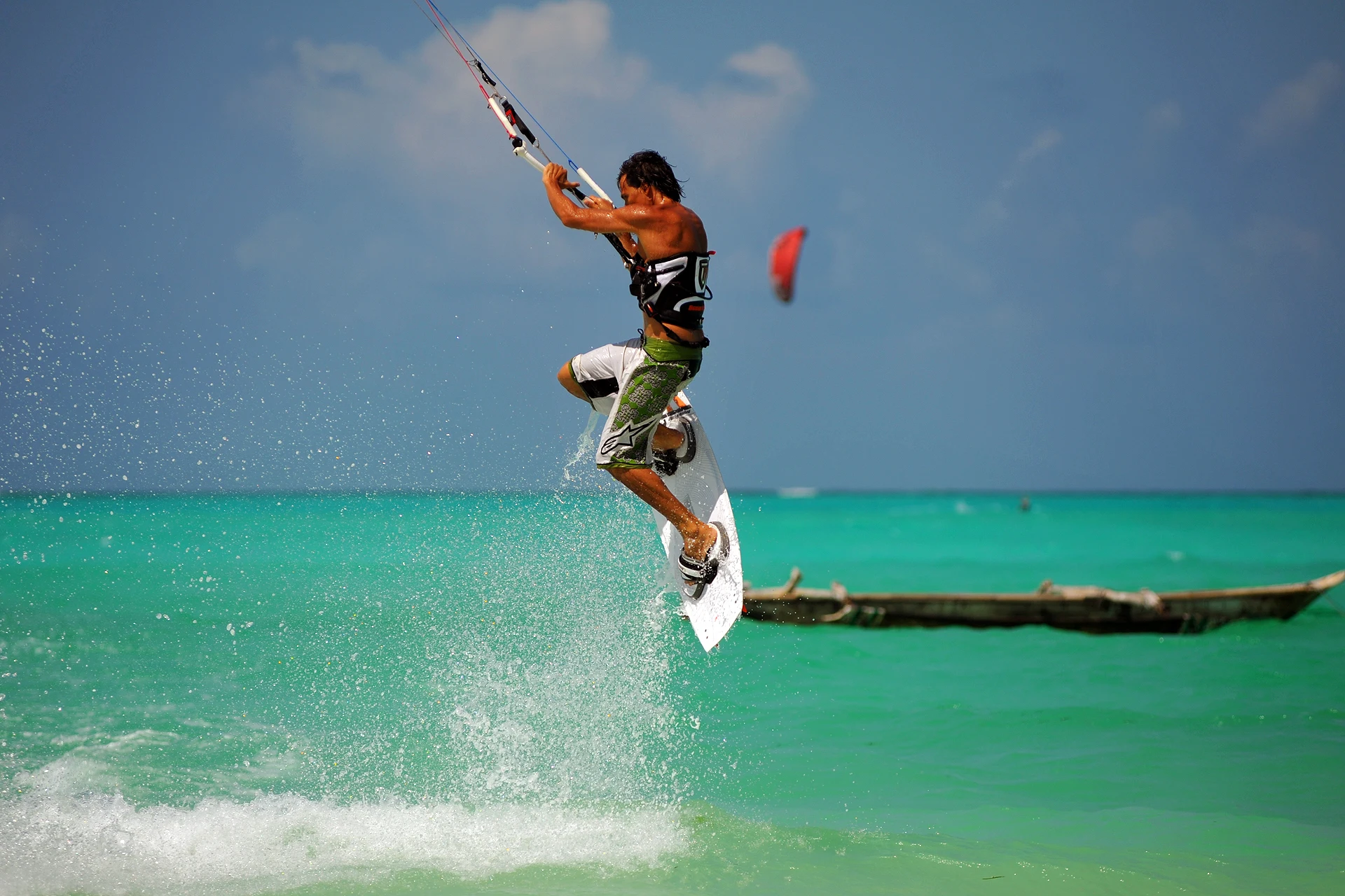 A kitesurfer jumping above turquoise water past a moored outrigger boat in Zanzibar