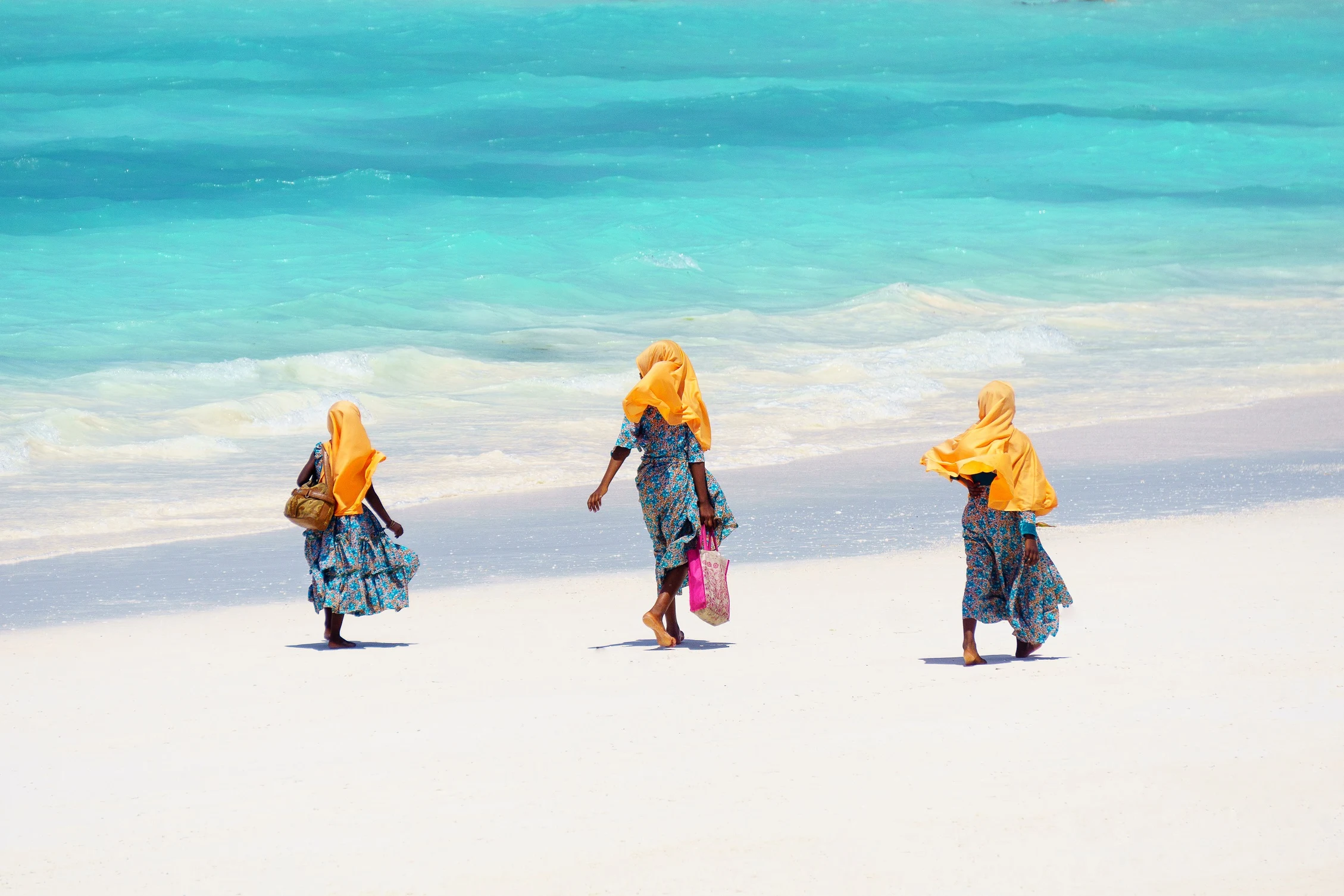 Three women in bright headscarves walking along a white beach