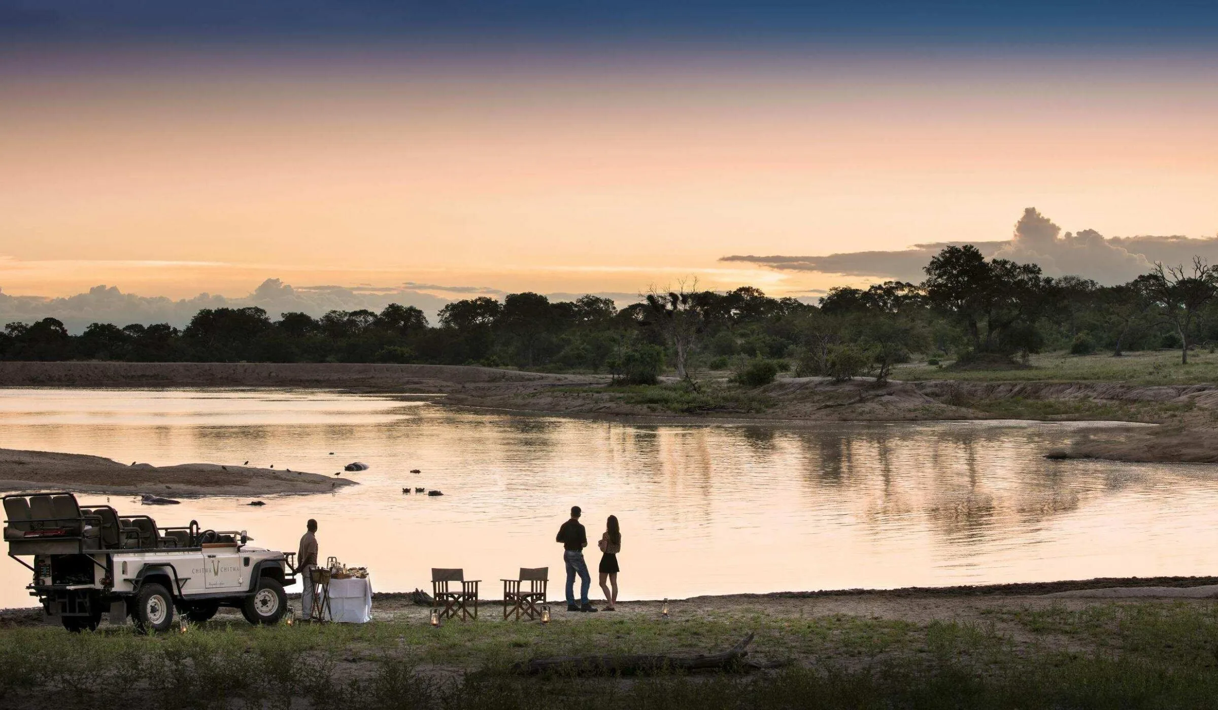 A couple beside a river at sunset next to their safari vehicle
