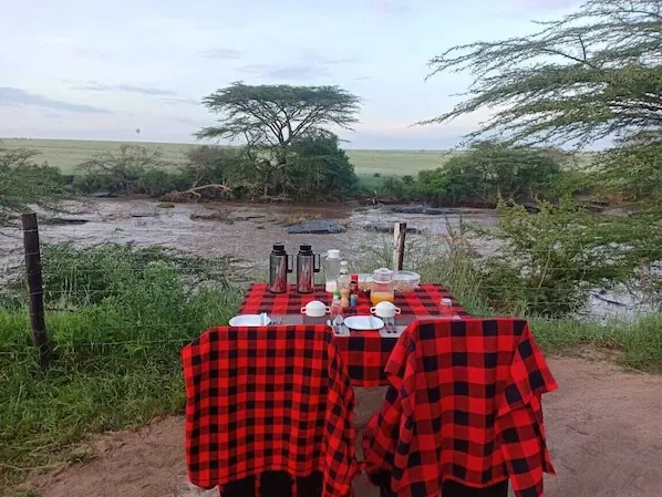 A dining table set up outdoors with a view of a river and trees in the background. - Mara Duma Bush Camp (Maasai Mara)